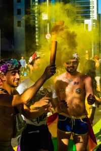 Photo of three male presenting people celebrating LGBTQ+ pride wearing rainbow flower crowns, body glitter, and flags. They're smiling widely waving yellow smoke wands.